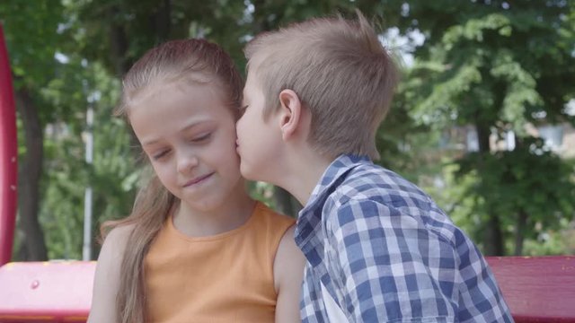 Portrait of adorable blond boy kissing cheek of a pretty girl sitting on the swing on the playground. The girl is shy. A couple of happy children. Funny kids in love