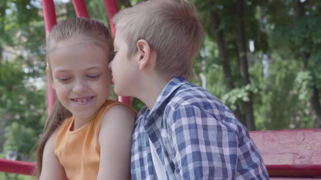 Portrait of cute blond boy kissing cheek of a pretty girl sitting on the swing on the playground. The girl is shy. A couple of happy children. Funny kids in love