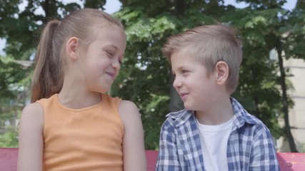 Portrait of cute blond boy and pretty girl sitting on the swing on the playground. Couple of happy children. Funny kids in love outdoors.
