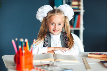Portrait of a beautiful girl in the classroom. Little schoolgirl with white bows sitting at the table and studying. Education and school concept-little girl with many books, pencils at school.