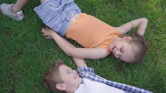 Two funny kids lying on the grass in the park smiling to each other. The girl taking hand of the boy. A couple of happy children. Funny carefree kids in love. Top view