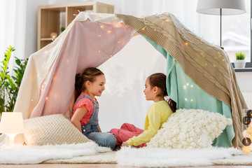 childhood, hygge and friendship concept - happy girls lying in kids tent and talking at home