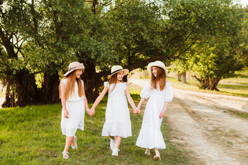 Naklejka na ściany i meble Three girls in white dresses walk in nature in the summer. Children's pastime during the summer holidays.