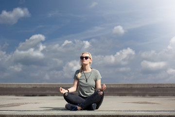 Blonde urban woman in dark jeans and sneakers, sitting in the middle of pavement, taking a break and meditating, with beautiful blue sky with white clouds behind her, on a bright sunny day