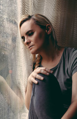 Blonde woman sitting by the rainy window, looking absently out, sad, lonely and depressed