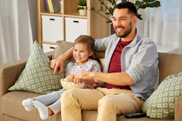 family, fatherhood and people concept - happy father and daughter with popcorn watching tv at home