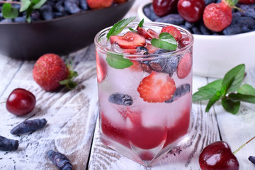 Lemonade with cherries, honeysuckles, strawberries, red syrup and ice in a glass topped with mint on the white table