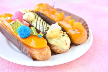 Delicious French eclairs with orange creative decor and colorful sugar raspberries on the white plate at pink textured background, selective focus. Traditional homemade profiteroles with orange icing
