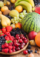 Fresh organic summer berries mix in wooden tray and exotic fruits on wooden background. Raspberries, strawberries, blueberries, blackberries and cherries. Watermelon, pear, pineapple, grapes, banana.