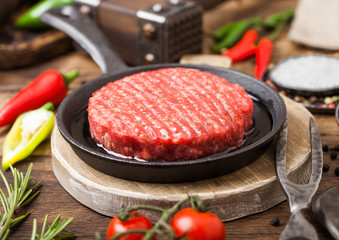 Fresh raw minced homemade farmers grill beef burgers on round chopping board and frying pan with spices and herbs and meat hatchet on wooden board. Oil, garlic and tomatoes.