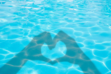 Shadows of hands forming a heart on blue swimming pool water background, summer concept