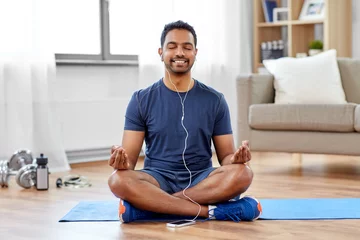 Poster fitness, meditation and healthy lifestyle concept - indian man in earphones listening to music on smartphone and meditating in lotus pose at home © Syda Productions