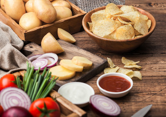 Fresh organic homemade potato crisps chips in wooden bowl with sour cream and red onions and spices on wooden table background. Fresh yellow potatoes with ketchup