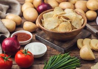 Fresh organic homemade potato crisps chips in wooden bowl with sour cream and red onions and spices on wooden table background. Fresh yellow potatoes with ketchup