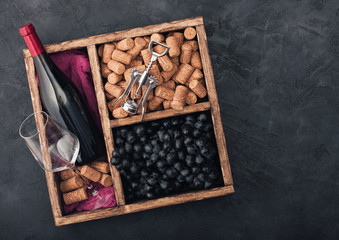 Bottle of red wine and empty glass with dark grapes with corks and corkscrew inside vintage wooden box on black stone background with red cloth. Top view