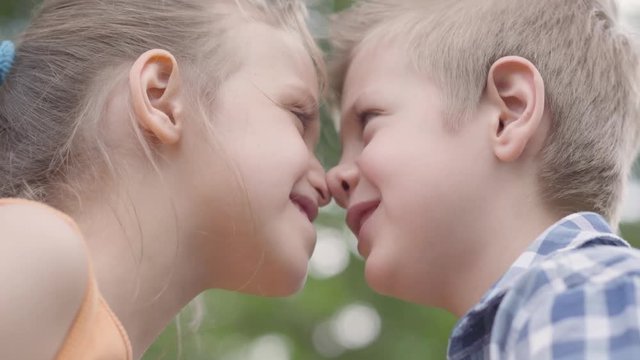 Close-up portrait of cute boy and girl sitting in the park, trying to rub their noses and having fun. A couple of happy children. Funny carefree kids in love outdoors.