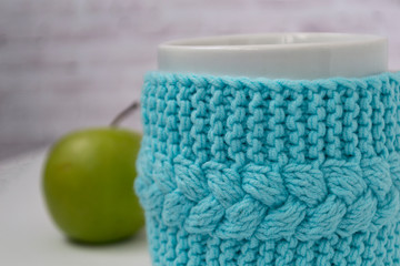 A cup in a knitted blue case with a green apple, fruit on a light background. The concept of comfort, relaxation, healthy food. Scandinavian style. There is a place for the test.