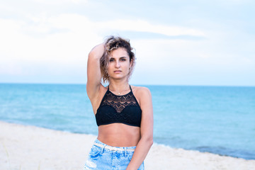 tanned beautiful young woman in denim shorts and swimsuit on the beach in clouds weather