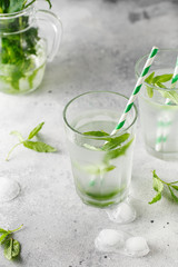 glasses of cold water with fresh mint leaves and ice cubes on grey concrete background.