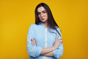 Cute young girl has upset look, curves lower lip, expresses dissatifaction and discontent, frowns face, isolated over yellow wall