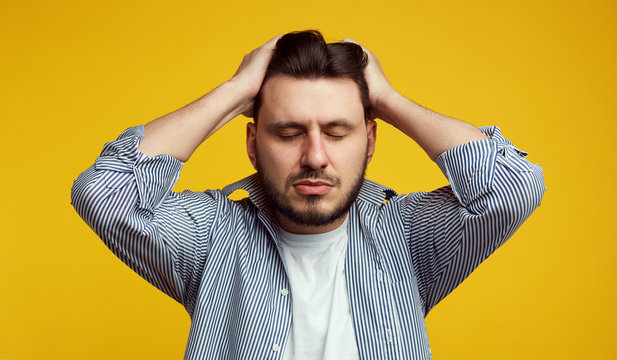 Upset young man keeps both hands on head, closes eyes, suffers from migraine, has sad fatigue facial expression wears casual shirt