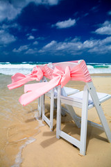 Chairs with pink ribbons blowing in the wind set up on a tropical beach for a wedding ceremony celebration with waves crashing on the shore and a blue sky