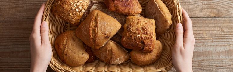 top view of woman holding fresh homemade buns in wicker basket above wooden table, panoramic shot