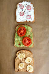 Three different toasts: cream cheese with radish and basil, avocado with cherry tomato and smoked paprika, peanut butter with banana and cinnamon. Top view, brown background.