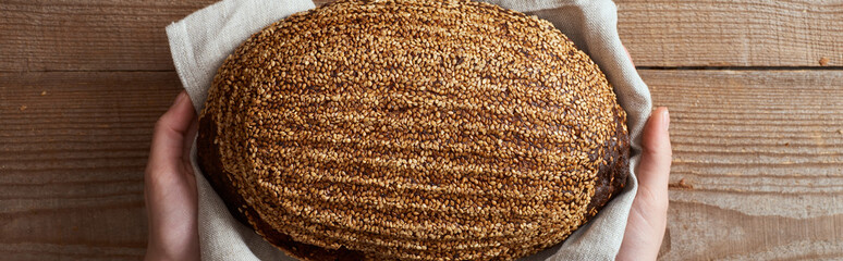 partial view of woman holding loaf of brown bread in towel above wooden surface, panoramic shot