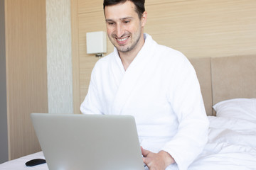 Beautiful young man using laptop while sitting on a bed.