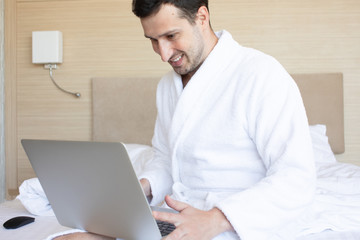 Beautiful young man using laptop while sitting on a bed.