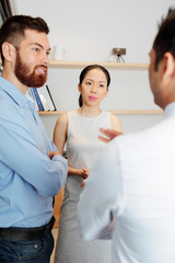 Bearded young businessman standing together with Asian businesswoman and they listening to their colleague while standing at office