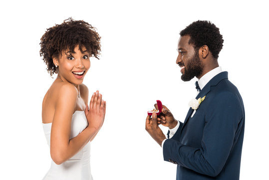 cheerful african american man holding box with ring while making proposal to surprised woman isolated on white