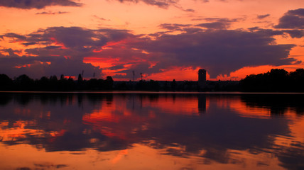 Amazing orange sunset over the horizon with a silhouette of isolated tower. Darkness and dramatic sky in beautiful landscape. Reflection and symmetry of the clouds. 