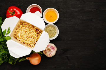 Dry noodles in paper box with greenery, vegetables and spices on a black table, top view, text space