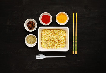 Instant noodles in container with plastic fork, chopsticks and spices on a black wooden table, top view