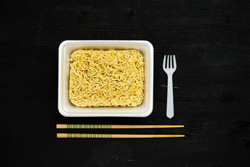 Instant noodles in container with plastic fork and chopsticks on a black wooden table, top view