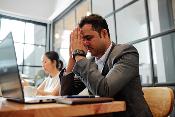 Indian businessman sitting at the table with laptop with is eyes closed and resting while businesswoman working near by him at office