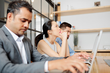 Asian young businesswoman sitting at the table and thinking about something while her colleagues working on laptop computers at office