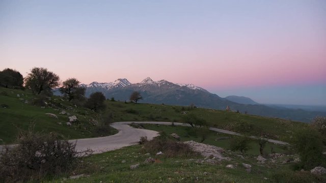 Night to day timelapse on a mountain road. View of high mountains with snow and green grass with the sunlight moving the shadows in the early morning.