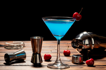 Colorful cocktail blue martini recipe with red cherry and bartender accessories on the wooden table in black background