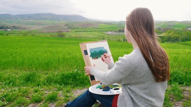 A talanted woman sits on the field and draws using a brush and paletes.
