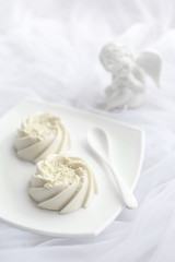 Light airy dessert panna cotta mousse with white chocolate and vanilla on top, on white ceramic plate and ceramic spoon and porcelain angel statuette. Monochrome