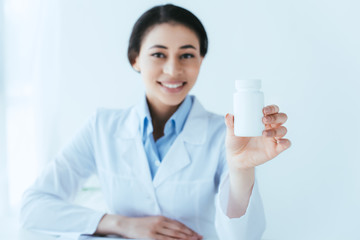selective focus of cheerful latin doctor holding pills container and smiling at camera