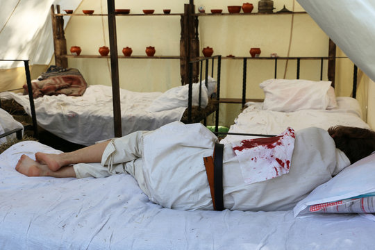 Wounded Soldier Lying On The Bed In A Field Military Hospital. Reconstruction Of Military Medicine Of The 19th Century