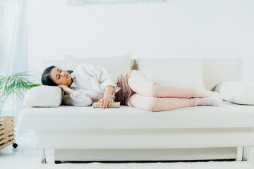 tired latin woman sleeping with book while lying on sofa at home