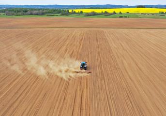Aerial perspective view on blue tractor pulling a plow, preparing a soil for seed sowing, tractor making dirt cloud. Landmark on meadow and rapeseed field