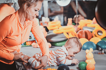 Young happy mom is plaing with her newborn baby at masseur cabinet full of special treatment toys. - obrazy, fototapety, plakaty