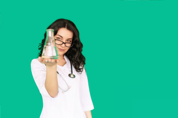 Girl nurse with glasses, white coat, with a stethoscope around his neck, in the laboratory, holding a flask, examines the contents, on a green background screen