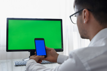Engineer, Constructor, Designer in Glasses Working on a Personal Computer with a Green Screen on Monitor which has Chroma Key Great for Mockup Template. Using Smartphone or Tablet with Blue Screen